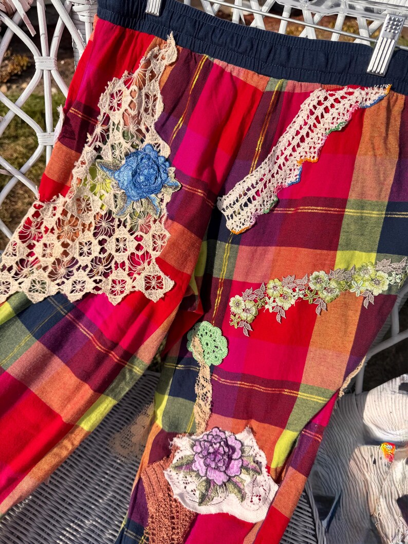 May include: Colorful plaid pants with decorative lace and floral patches. The pants feature a mix of red, pink, yellow, and blue plaid, with various lace and embroidered floral accents. The waistband is a dark blue.