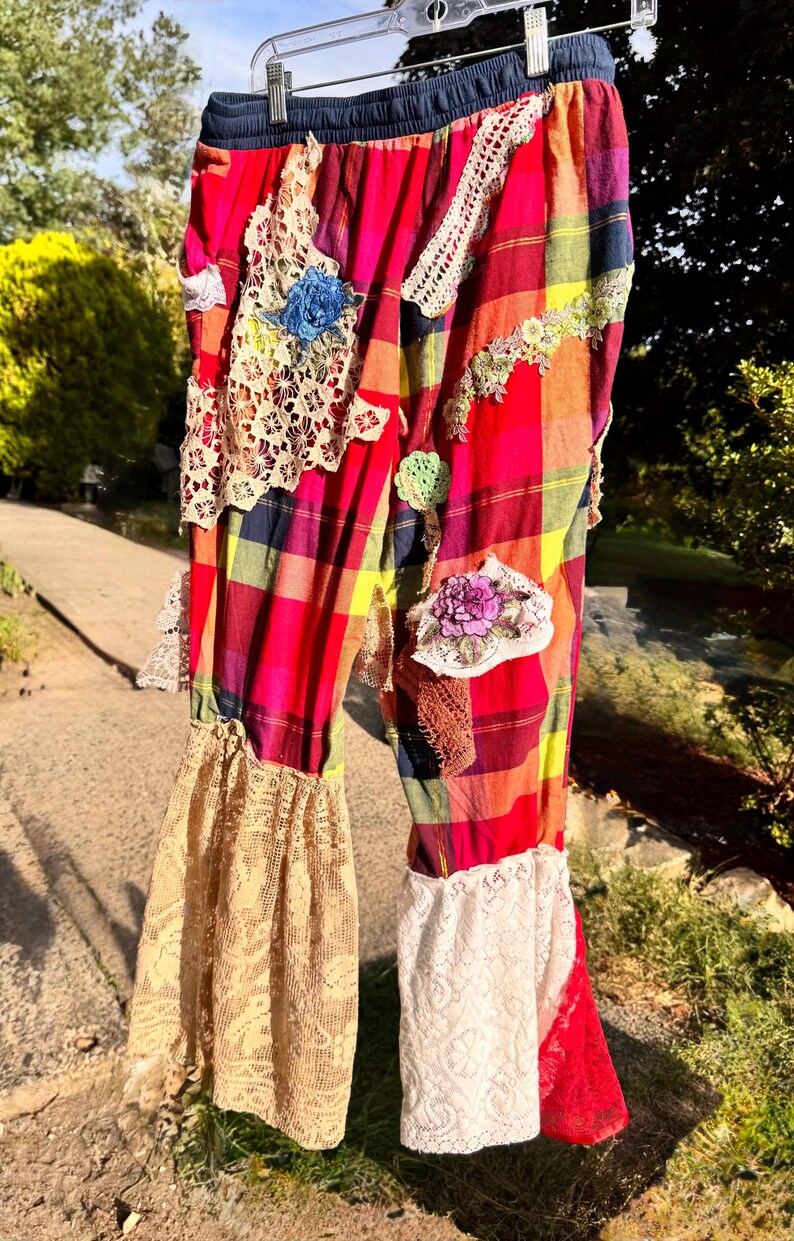 May include: Colorful plaid pants with lace and floral embellishments. The pants feature a red, yellow, and blue plaid pattern, with patches of lace and embroidered flowers. The pants have a drawstring waist and flared lace cuffs.