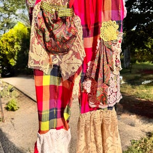 May include: Colorful plaid pants with a drawstring waist, adorned with lace, floral embroidery, and paisley fabric patches. The pants feature a mix of red, yellow, and blue hues, with a bohemian style. The pants have a flared bottom and are hanging outside.