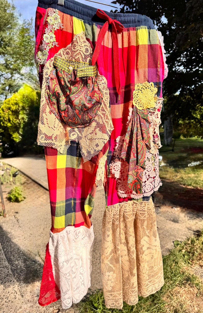 May include: Colorful plaid pants with a drawstring waist, adorned with lace, floral embroidery, and paisley fabric patches. The pants feature a mix of red, yellow, and blue hues, with a bohemian style. The pants have a flared bottom and are hanging outside.