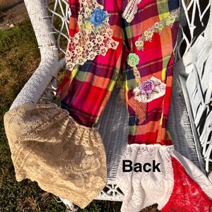 May include: Colorful plaid pants with lace and floral appliqu&eacute;s. The pants feature a mix of red, pink, yellow, and blue plaid, with decorative lace patches and floral accents. The bottom of the pants have a flared, lace trim. The word "Back" is visible on the pants.