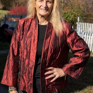 Może przedstawiać: Czerwona kurtka z teksturowanym wzorem. Kurtka jest otwarta, odsłaniając czarny top pod spodem. Osoba ma na sobie ciemne dżinsy i biżuterię.