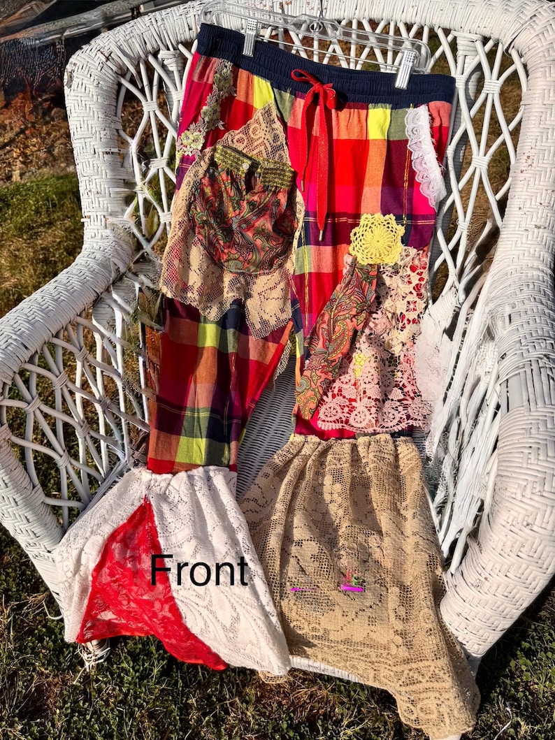 May include: Colorful patchwork pants with a red drawstring waist. The pants feature a mix of plaid, paisley, and lace fabrics in shades of red, yellow, and beige. A yellow crocheted flower is sewn onto the front. The word "Front" is visible on the pants.