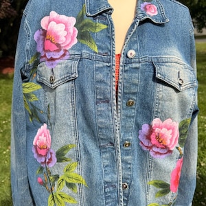 May include: A blue denim jacket featuring hand-painted pink and purple floral designs with green leaves. The jacket includes a classic collar, a button-down front with metallic buttons, and two chest pockets with buttoned flaps on the front panels.