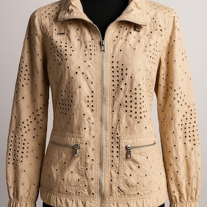 May include: A beige zip-up jacket featuring an all-over eyelet lace pattern with circular cutouts. The jacket includes a stand-up collar, long sleeves with elasticized cuffs, and two front pockets with silver-toned metal zippers and matching hardware.