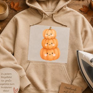 Könnte beinhalten: Ein beigefarbener Kapuzenpullover mit einer zentralen Grafik aus drei gestapelten orangefarbenen Kürbissen mit lächelnden Gesichtern und rosigen Wangen. Der Hoodie hat lange Ärmel, eine Kängurutasche und passende Kordelzüge am Ausschnitt.