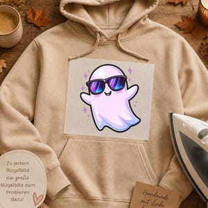 May include: A beige hooded sweatshirt featuring a central graphic of a light pink ghost wearing dark blue sunglasses with purple lenses. The ghost is surrounded by small purple star accents. The hoodie has a front kangaroo pocket and long sleeves.