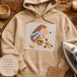 Könnte beinhalten: Ein beigefarbener Kapuzenpullover mit einer Grafik eines Igels, der einen blau-weiß gestreiften Schal und gelbe Stiefel trägt und einen Korb mit Kastanien hält. Das Motiv zeigt verstreute Herbstblätter und Kastanien auf hellem Grund.