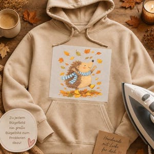 Könnte beinhalten: Ein beigefarbener Kapuzenpullover mit einer zentralen Grafik eines Zeichentrick-Igels, der einen blau-weiß gestreiften Schal trägt und von herbstlichen Blättern umgeben ist. Der Hoodie verfügt über eine Kängurutasche und passende Kordelzüge.