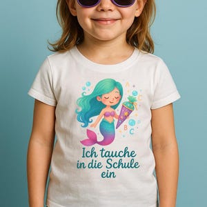 Könnte beinhalten: Ein weißes T-Shirt mit einer Meerjungfrauen-Illustration, die einen bunten Schultüten hält. Der Text auf dem Shirt lautet "Ich tauche in die Schule ein".
