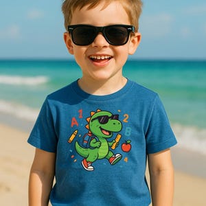 Könnte beinhalten: Ein kleiner Junge trägt eine Sonnenbrille und ein blaues T-Shirt mit einem Comic-Dinosaurier, der eine Sonnenbrille und einen Rucksack trägt. Der Dinosaurier hält ein Lineal und einen Apfel. Das T-Shirt hat die Buchstaben A, B, 1, 2 und 4 aufgedruckt.
