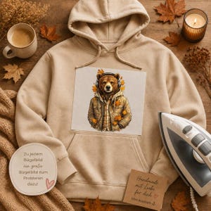 Könnte beinhalten: Ein beigefarbener Kapuzenpullover mit einem zentralen Aufdruck eines braunen Bären, der Kopfhörer und eine beigefarbene Jacke trägt, umgeben von orangefarbenen Herbstblättern. Der Hoodie enthält eine runde und eine quadratische Karte mit deutschem Text.