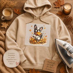 Könnte beinhalten: Ein beigefarbener Kapuzenpullover mit einem Grafikdruck einer Französischen Bulldogge, die einen roten Schal trägt und ein gelbes Herbstblatt im Maul hält, umgeben von herabgefallenen Blättern. Der Hoodie hat eine Kängurutasche und passende Kordeln.