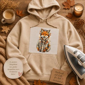 Könnte beinhalten: Ein beigefarbener Kapuzenpullover mit einem zentralen Aufdruck eines Fuchses, der Kopfhörer und eine Jacke trägt, umgeben von Herbstblättern. Der Hoodie verfügt über eine Kängurutasche und passende Kordeln, präsentiert mit einem kleinen Bügeleisen.