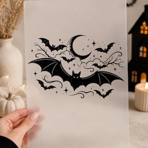 Könnte beinhalten: Ein durchscheinendes Blatt mit einer schwarzen Tuschezeichnung einer großen Fledermaus mit ausgebreiteten Flügeln, umgeben von kleineren Fledermäusen, einer Mondsichel, wirbelnden Wolken, kahlen Ästen und kleinen Sternen.