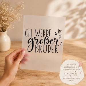 Puede incluir: Una hoja rectangular semitransparente con el texto impreso en negro "ICH WERDE großer BRUDER" acompañado de tres pequeños iconos de corazón. El diseño utiliza una combinación de tipografía sans-serif y cursiva sobre una superficie mate.