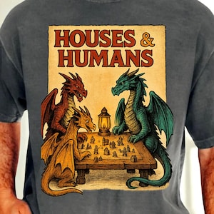 Könnte beinhalten: Dunkelgraues T-Shirt mit einer Grafik von drei Drachen, die ein Brettspiel spielen. Die Grafik enthält den Text "HOUSES & HUMANS" im Vintage-Stil. Die Drachen sind rot, gold und grün, mit einer Laterne auf dem Tisch.
