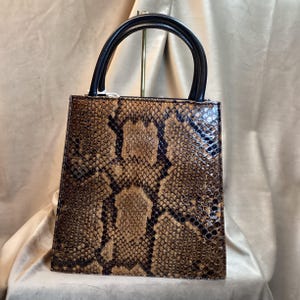 May include: A structured handbag with a snakeskin pattern in shades of brown and black. The bag has a rectangular shape with a black handle. The bag is made of a textured material.