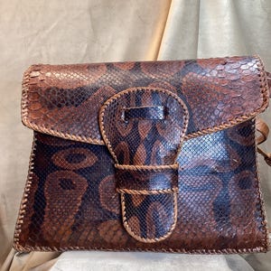 May include: A brown and black snakeskin shoulder bag with a flap closure. The bag features a unique clasp design and is stitched with a lighter brown thread. The bag has a textured surface and a strap.