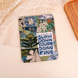 May include: A decorative rectangular item with a collage design. The design includes floral patterns, a dragonfly, and the text "SLOW DOWN YOURE DOING FINE" in blue and silver. A wicker basket is in the background.