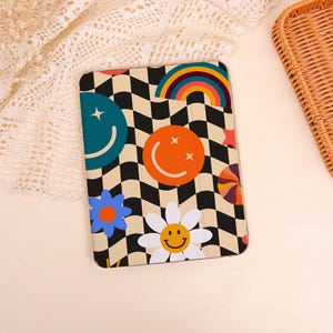 May include: A tablet case with a black and white checkered pattern, featuring smiley faces, flowers, a rainbow, and stars. The case has a beige background and a retro, groovy aesthetic.