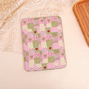 May include: A rectangular tablet case featuring a patchwork pattern of pink, olive green, and white squares. The design includes various geometric motifs such as stripes, zigzags, and dots, interspersed with small solid green and red heart icons.