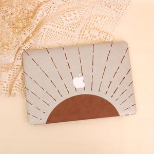 May include: A laptop case featuring a sunburst design. The case has a light beige background with a brown half-circle at the bottom and brown lines radiating outwards. The Apple logo is visible in the center. The case is on a beige surface.