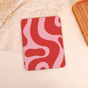 Peut inclure: Une coque de tablette rouge avec un motif ondulé rose. L'étui est rectangulaire et présente une surface lisse. Le motif abstrait évoque un style rétro. L'étui est présenté sur une surface de couleur claire.
