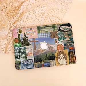 May include: A decorative laptop case featuring a collage of images and text. The design includes a mountain scene with an Apple logo, a wooden cross, and various nature-themed stickers. The case has a glossy finish and a rectangular shape.