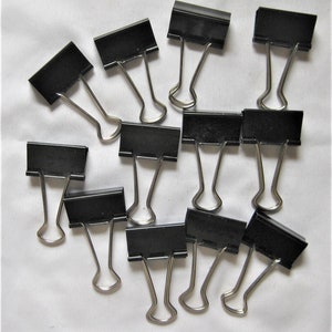Peut inclure: Un groupe de 12 clips de reliure noirs, tous de même taille et de même style, sont éparpillés sur une surface blanche. Les clips sont en métal et ont une finition argentée.