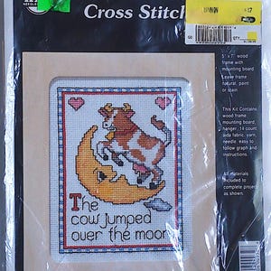 May include: A counted cross stitch kit featuring a brown and white cow jumping over a yellow moon. The kit includes a 5x7 inch wooden frame, mounting board, fabric, yarn, needle, and instructions. The kit is titled "The cow jumped over the moon".