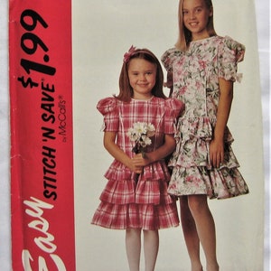 Può includere: Un cartamodello per un vestito da bambina. Il cartamodello è per le taglie 7-8, 10-12. Il cartamodello si chiama "Easy Stitch 'n Save" di McCall's. Il cartamodello mostra due bambine che indossano il vestito. Una bambina indossa un vestito a quadri rosa e bianco e l'altra bambina indossa un vestito a fiori.