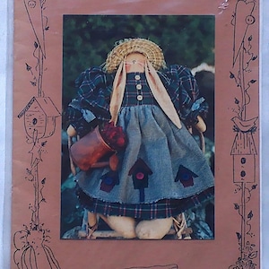 May include: A pattern for a 13-inch gardening bunny doll named Abigail. The doll is wearing a blue and green plaid dress with a straw hat and holding a watering can. The pattern is from Wendy's Cottonthread Cottage.