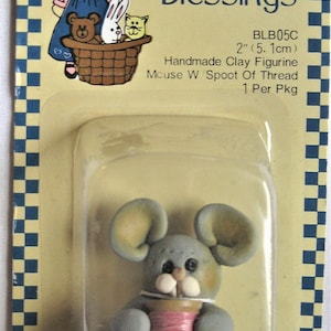 May include: A small gray clay figurine of a mouse holding a spool of pink thread. The figurine is packaged on a card with the text "Little Blessings" and "BLB05C 2" (5.1cm) Handmade Clay Figurine Mouse W/Spoot Of Thread 1 Per Pkg".