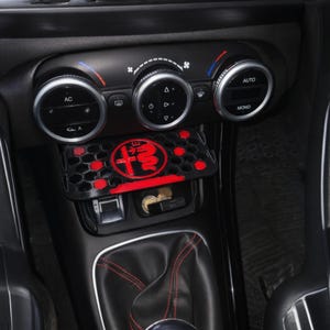 以下が含まれることがあります： 黒い車のインテリア。赤と黒の収納コンパートメントにはアルファロメオのロゴが入っています。コンパートメントはエアコンコントロールの下に配置されています。ギアシフトには赤いステッチが施されています。
