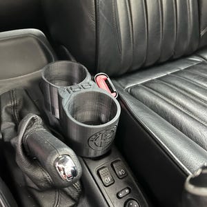 以下が含まれることがあります： 車の内装用のカスタムメイド3Dプリントカップホルダー。 ホルダーはグレーで、カップ用の2つの円形の開口部があり、「156」の文字とアルファロメオのロゴが付いています。 ギアシフトとダッシュボードコントロールの近くに配置されています。
