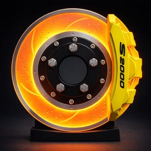 Op de afbeelding: Een decoratieve, verlichte remschijf en remklauw. De schijf gloeit oranje met een zilveren rand en zwart midden. De gele remklauw is voorzien van de tekst "S2000" in zwart. Het stuk is gemonteerd op een zwarte basis.