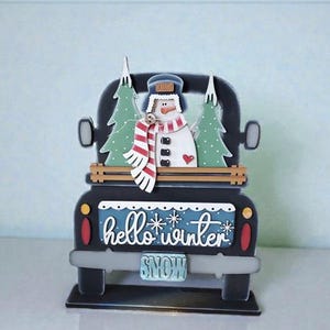 May include: A black wooden truck decoration with a snowman and two green Christmas trees in the bed. The truck has the words "hello winter" and "snow" on it. The snowman wears a hat and a red and white striped scarf.