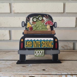 May include: A black wooden truck decoration with a frog wearing glasses in the bed. The truck has the words "Hop Into Spring" and "Ribbit" on it. The frog is surrounded by mushrooms and plants.