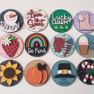 May include: A collection of twelve round wooden decorative signs with various seasonal and holiday-themed designs. The signs feature images of a snowman, strawberry, rainbow, pumpkin, and candy canes, among others. Each sign has a unique color scheme and text.