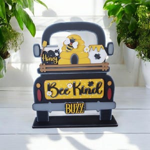 May include: A wooden decorative truck with a bee theme. The truck is black with a yellow panel that reads "Bee Kind" and "Buzz". It features a beehive, honey jars, and a bee. The truck is a charming addition to any home decor.