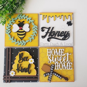 May include: Four wooden signs with a bee theme. The first sign has a bee with wings and a wreath of green leaves on a yellow background. The second sign has the word "Honey" in black script with a dripping honey design on a yellow background. The third sign has a beehive with the words "Bee Kind" on a black and white background. The fourth sign has a honeycomb pattern with the words "Home Sweet Home" on a yellow background.