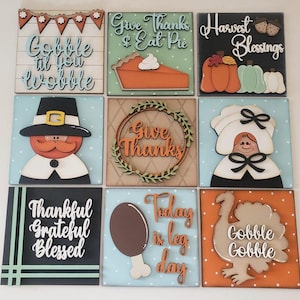 May include: Nine square wooden Thanksgiving decorations. Each square features a different design, including pumpkins, a turkey, a pilgrim, and phrases like "Gobble til you wobble" and "Give Thanks & Eat Pie."