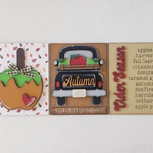 May include: Three wooden fall-themed signs. One features a caramel apple with a green bow and heart. The center sign shows a black truck with the word "Autumn". The third sign says "Cider Season" with a list of fall items.