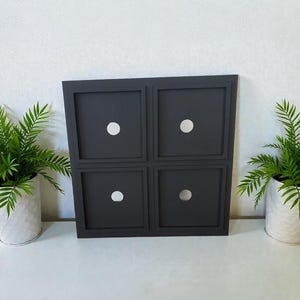 May include: A square, dark gray wall-mounted display panel featuring four recessed square compartments, each with a single circular cutout in the center. The panel is constructed with a flat frame and a smooth, matte finish throughout the surface.