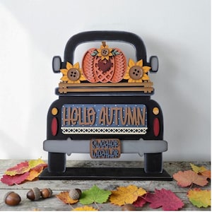 May include: A black wooden truck cutout featuring a pumpkin with a heart and sunflowers in the bed. The truck has a blue sign with white polka dots that reads HELLO AUTUMN and a license plate that reads SWEATER WEATHER in blue lettering.