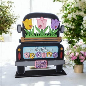 May include: A black wooden truck decoration with three colorful tulips in the bed. The truck has the words "hello SPRING" and "FLOWER MARKET" on it. The tulips are yellow, pink, and purple.