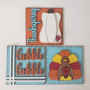 May include: Three wooden signs with Thanksgiving themes. The first sign is orange with blue text that says "Thanksgiving". The second sign is blue with white polka dots and orange text that says "Gobble Gobble". The third sign is blue with white polka dots and features a brown turkey with red, orange, and yellow feathers. The turkey has a red heart with the text "Gobble 'til you wobble" on it.