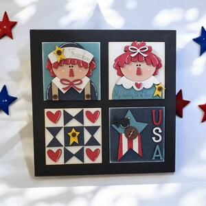 May include: A framed artwork featuring four square panels with patriotic themes. The top panels depict stylized figures with the word "Freedom" and the phrase "My country, 'tis of thee." The bottom panels display a quilt pattern and a star with "USA" lettering.