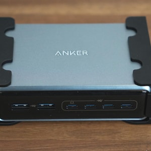 May include: A silver and black Anker docking station with multiple ports. The device has the brand name "ANKER" printed on the top. The docking station is rectangular with black brackets on the sides.